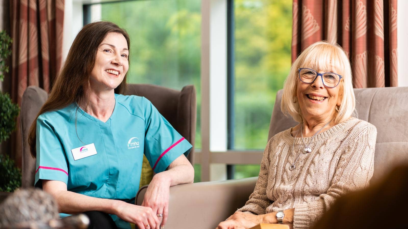 Two women sit in a well-lit room with large windows in the background. The woman on the left, likely a caregiver, wears a teal uniform with a name tag and smiles warmly. The older woman on the right, with short blonde hair and glasses, holds a book