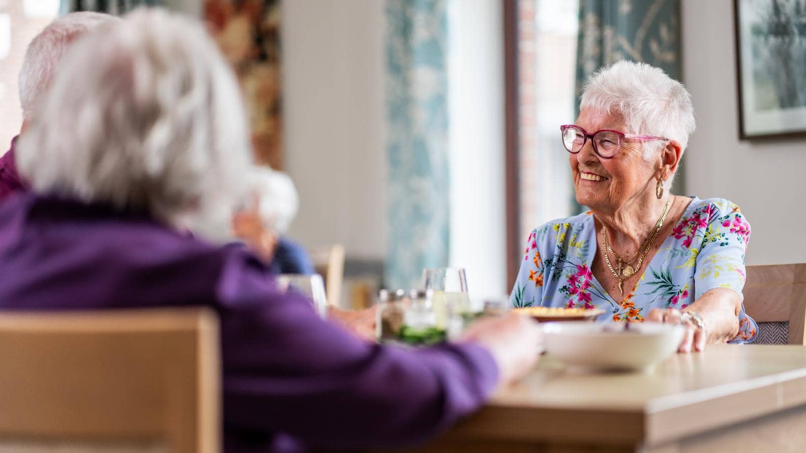 A cheerful elderly woman with short white hair and glasses sits at a dining table, wearing a floral dress and smiling as she converses. The bright dining area has soft lighting and decorative curtains.