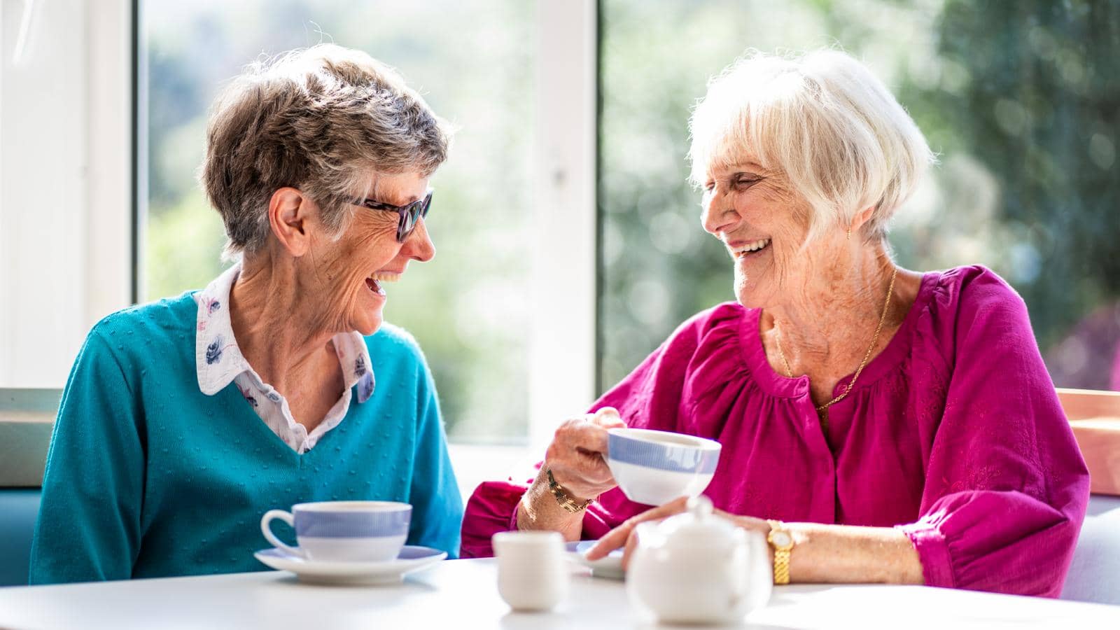  Two elderly women sit at a table, smiling and laughing together. One wears a blue sweater with a white collar, the other a bright pink top. A teacup is in hand, and a small teapot sits on the table