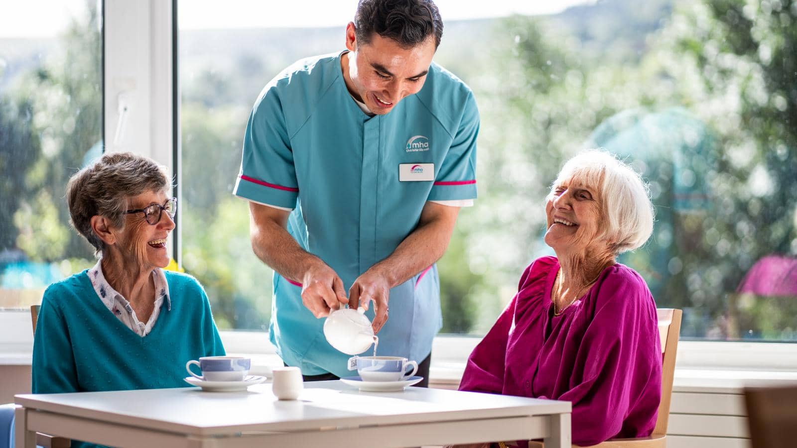 A male caregiver in a light blue uniform pours milk into a cup for two elderly women at a table. One woman, with gray hair and glasses, and the other, with white hair and a pink top, smile warmly.