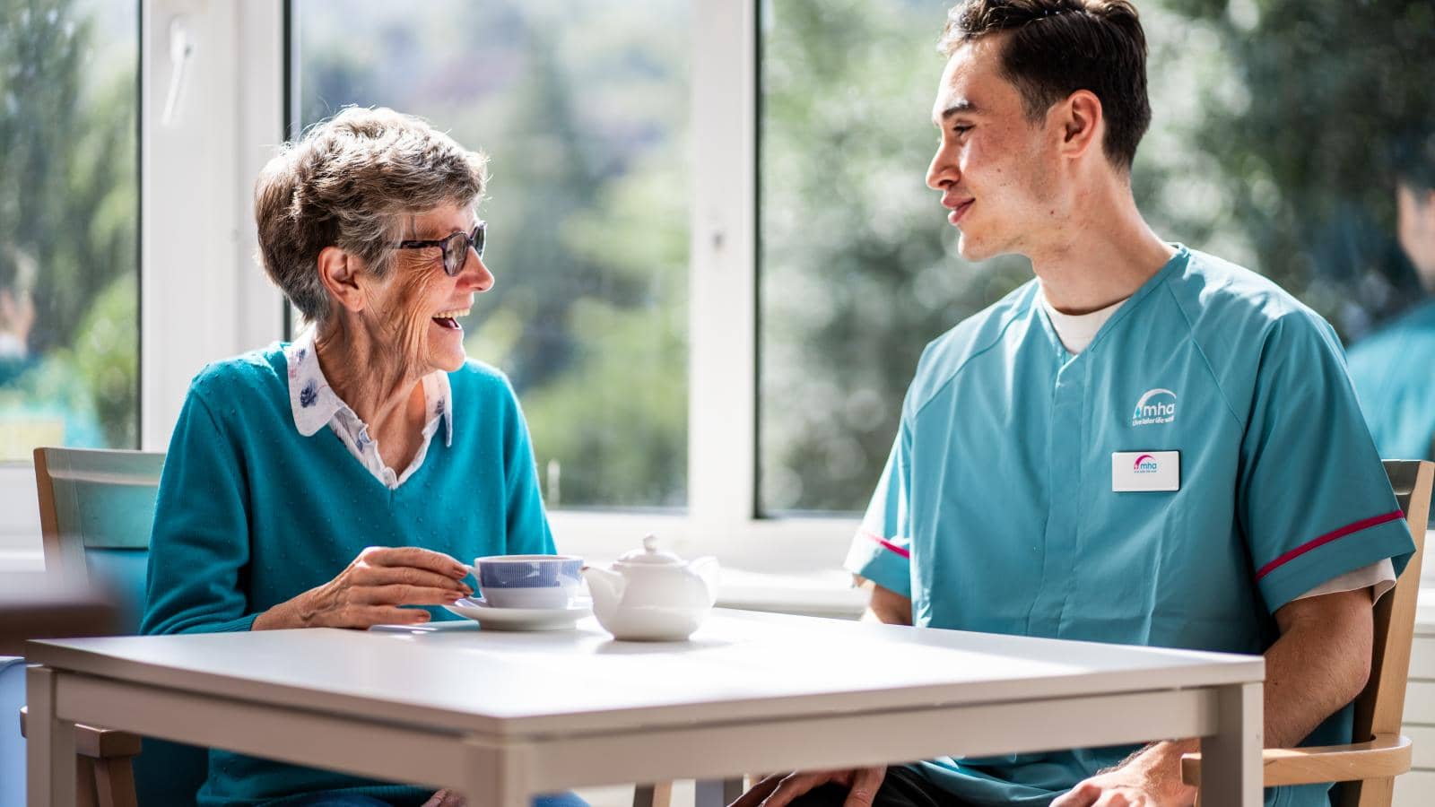 A caregiver and an elderly woman share a warm interaction at a table in a bright room with large windows allowing natural light. The elderly woman, wearing glasses, smiles as she converses, while the caregiver listens attentively.