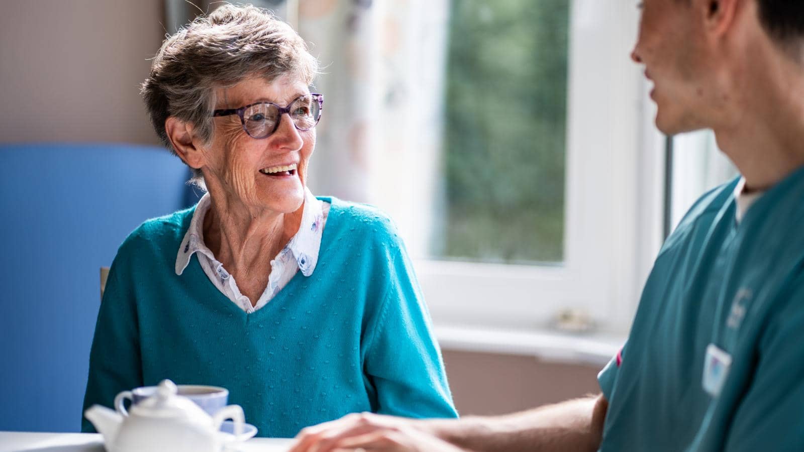 An elderly woman wearing glasses and a light blue sweater smiles at a caregiver in scrubs. They sit at a table with a teapot and cup, suggesting a cozy tea time.