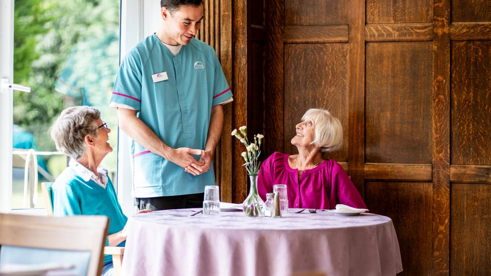 A young male caregiver interacts with two elderly women seated at a round table covered with a light purple tablecloth. One woman sits on the left, while the other, with short white hair, is on the right. They both appear engaged and happy