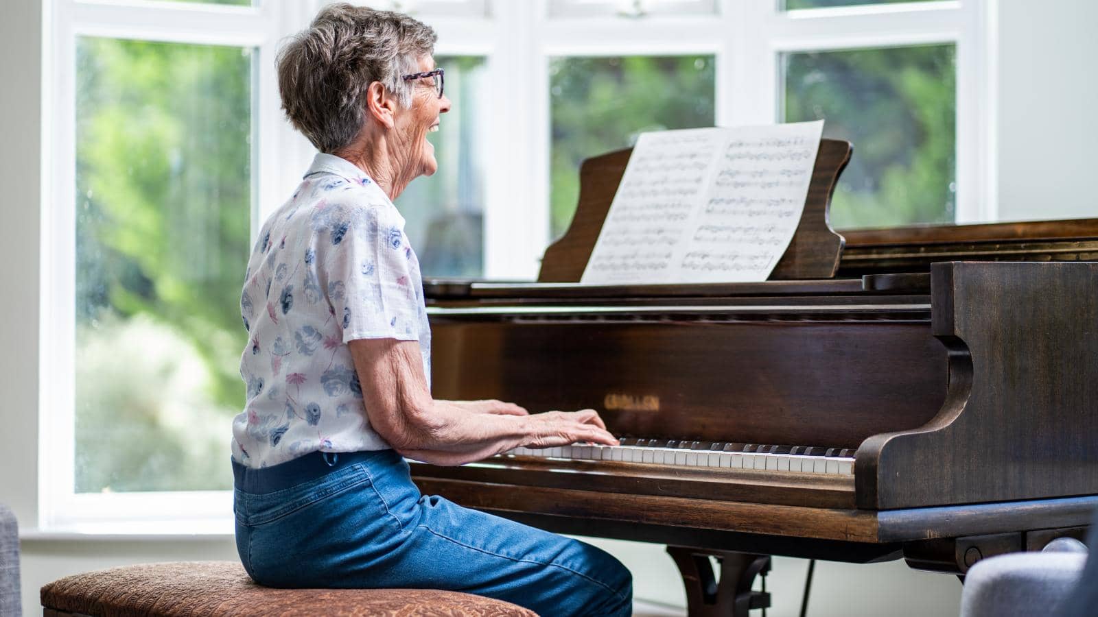 An elderly woman sits at a grand piano, playing with a joyful expression. The piano has a dark wood finish, with sheet music on the stand. The bright, airy setting features large windows letting in natural light and views of greenery.