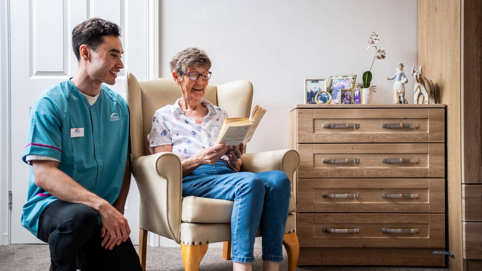 A caregiver, dressed in a teal uniform, is kneeling beside an elderly woman, who is seated in a comfortable armchair. She is reading a book and appears to be enjoying the moment. The room is well-lit and cozy, featuring a wooden dresser