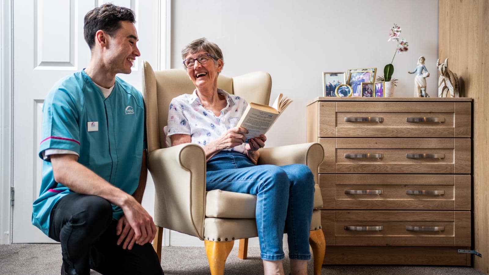 A friendly interaction between a carer and an elderly woman. The caregiver is kneeling beside a chair where the woman sits, smiling and holding a book