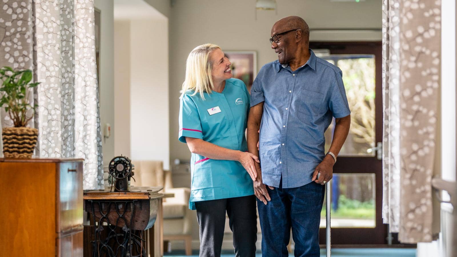 A caregiver in a light blue uniform smiles while walking with an elderly man who uses a cane. They move down a well-lit corridor with natural light streaming through windows. The care facility setting has comfortable furnishings and decor