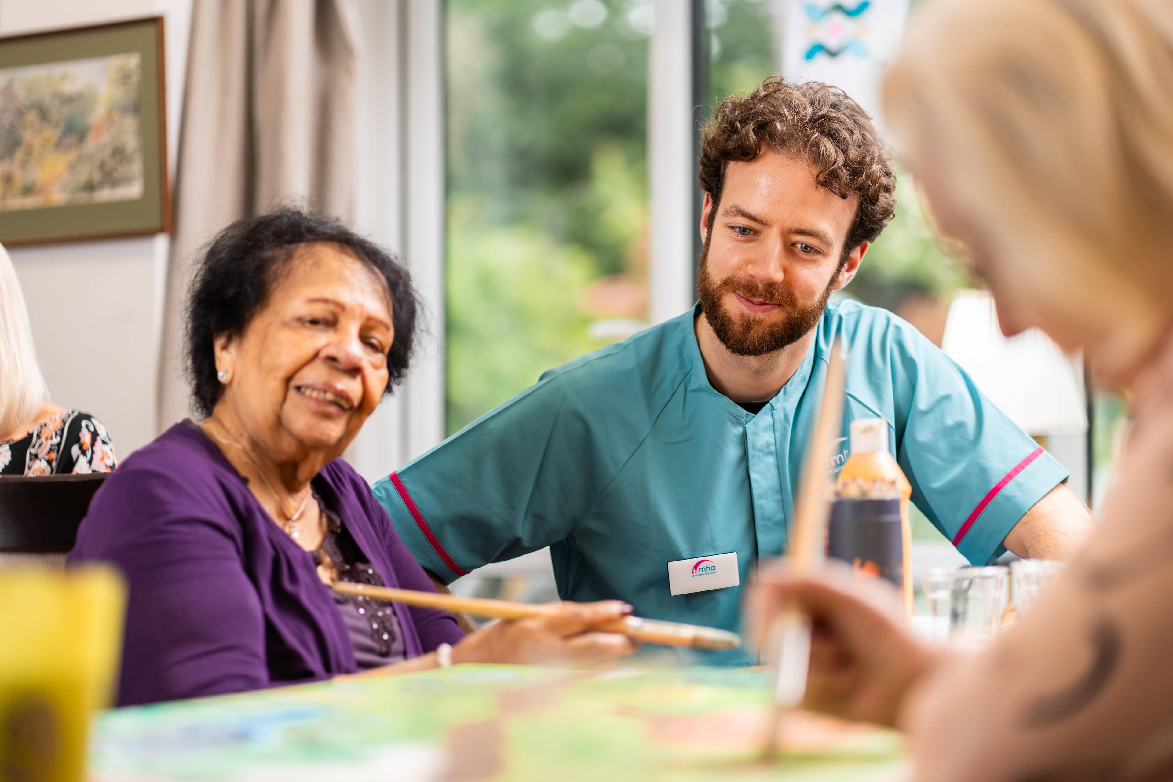 Female care home resident wearing a purple cardigan painting a picture whilst sat with a male care assistant