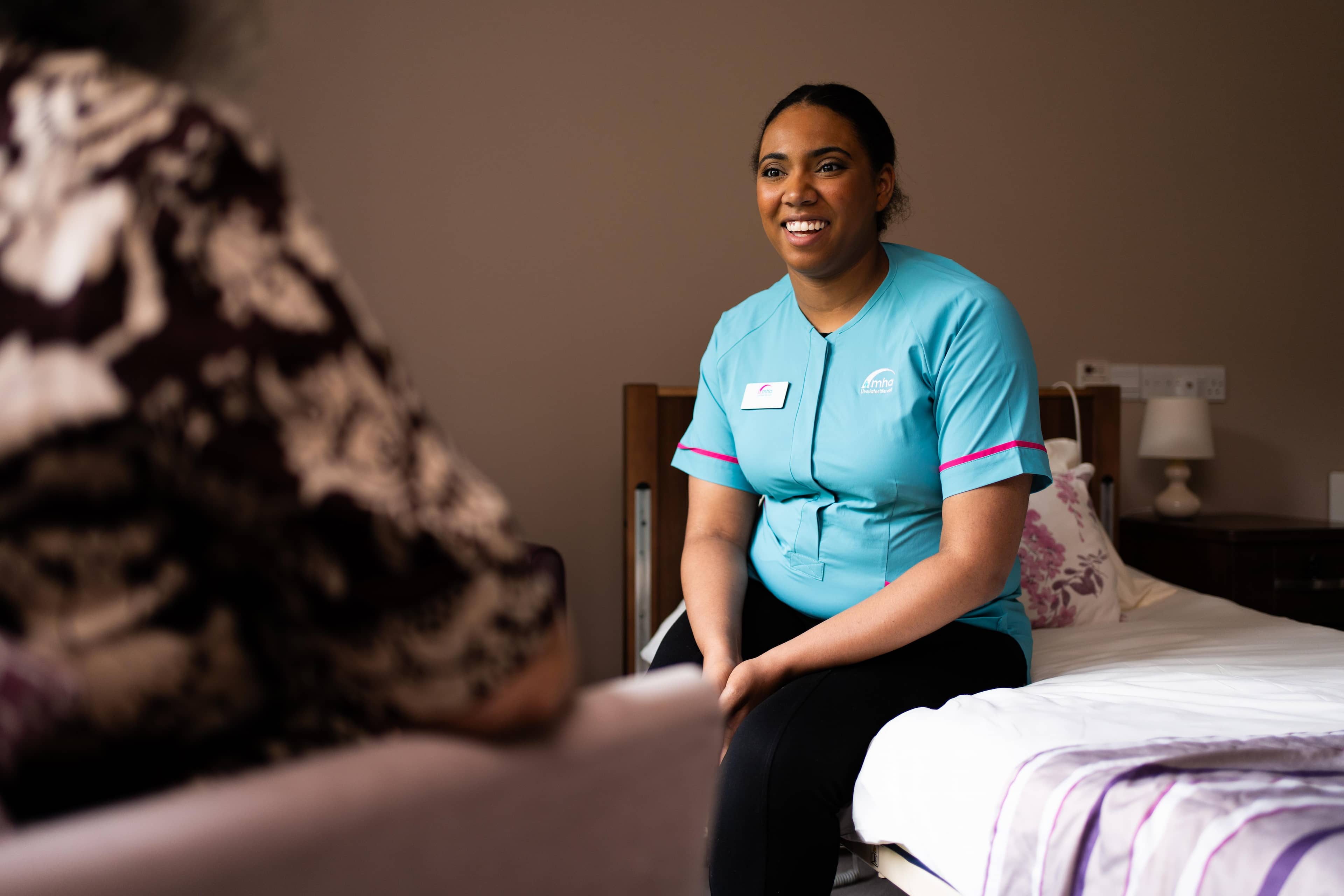 Female care assistant speaking to resident who is out of focus whilst sat on a bed