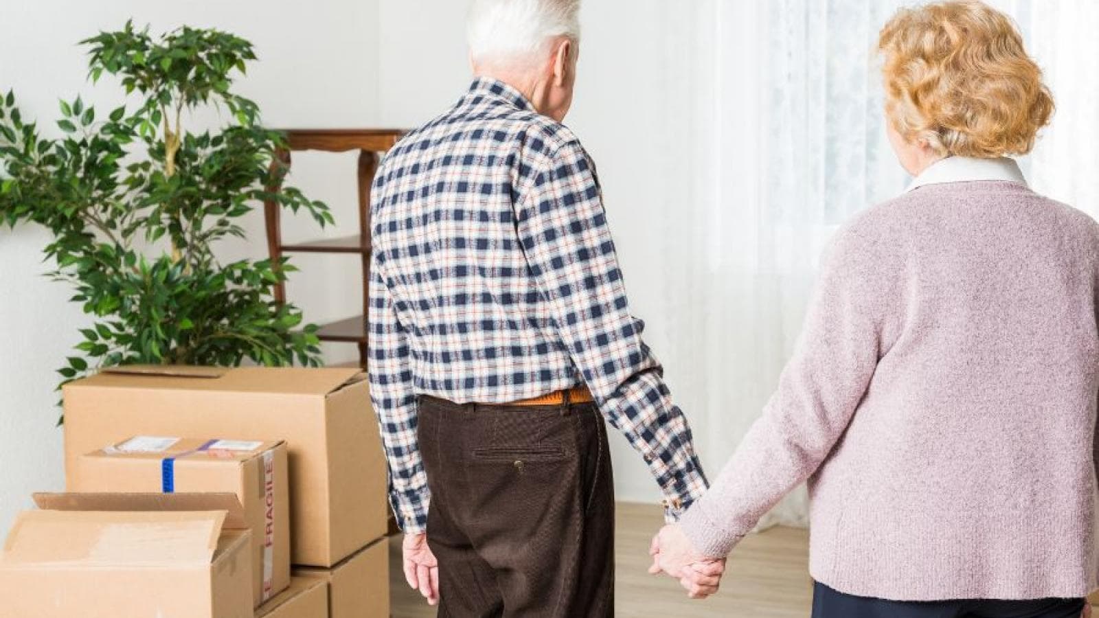 An older couple hold hands in their home looking at piles of cardboard boxes. 