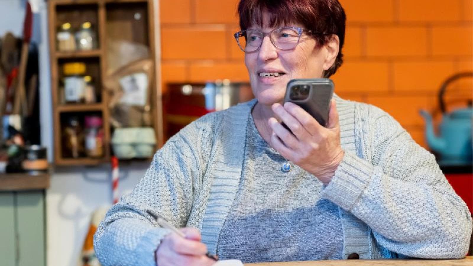 Woman sits at a table making notes whilst on the phone.