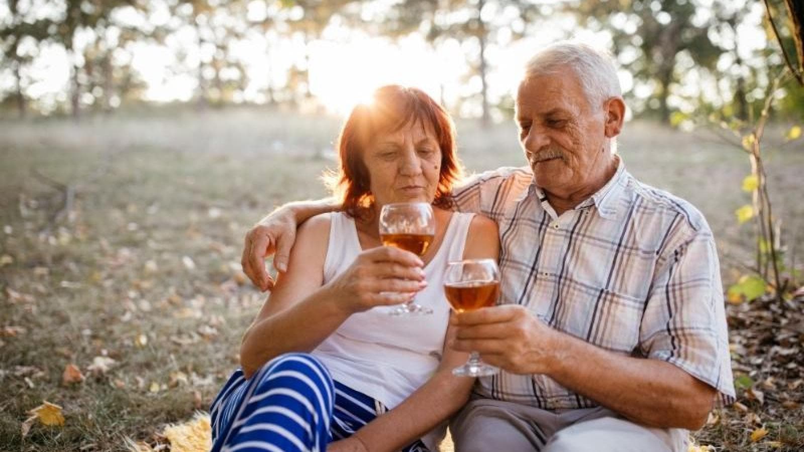 A couple sit in the sunshine with a wine glass each about to toast 