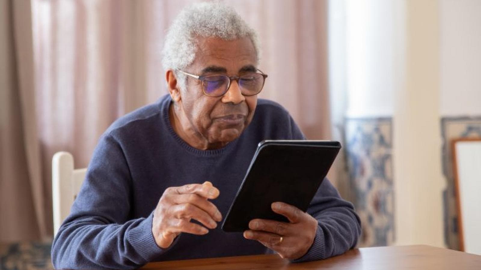 older man sitting at table looking at a tablet - stock image