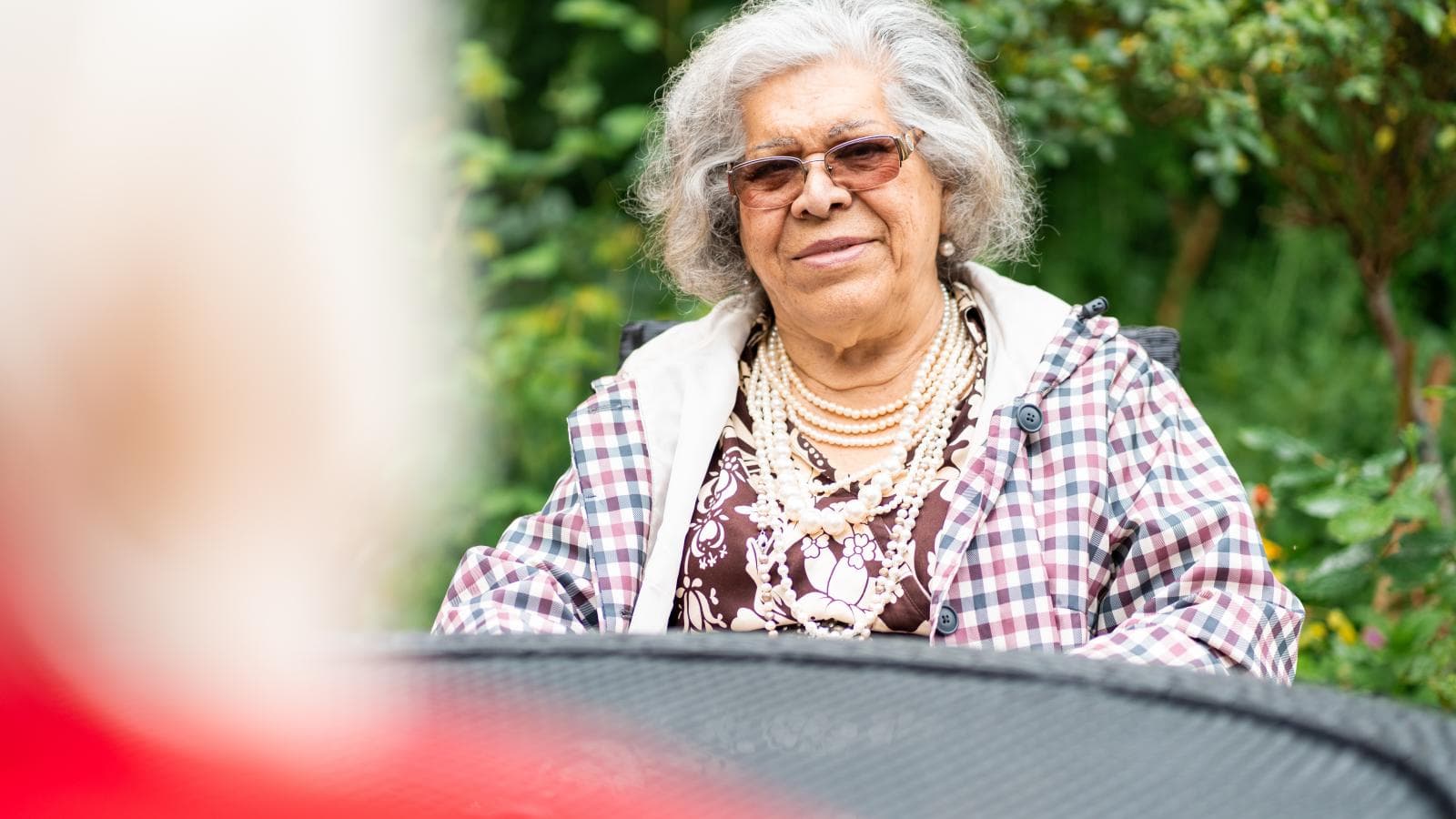 Older lady wearing glasses and a coat sat outside at a table smiling at someone who is out of focus
