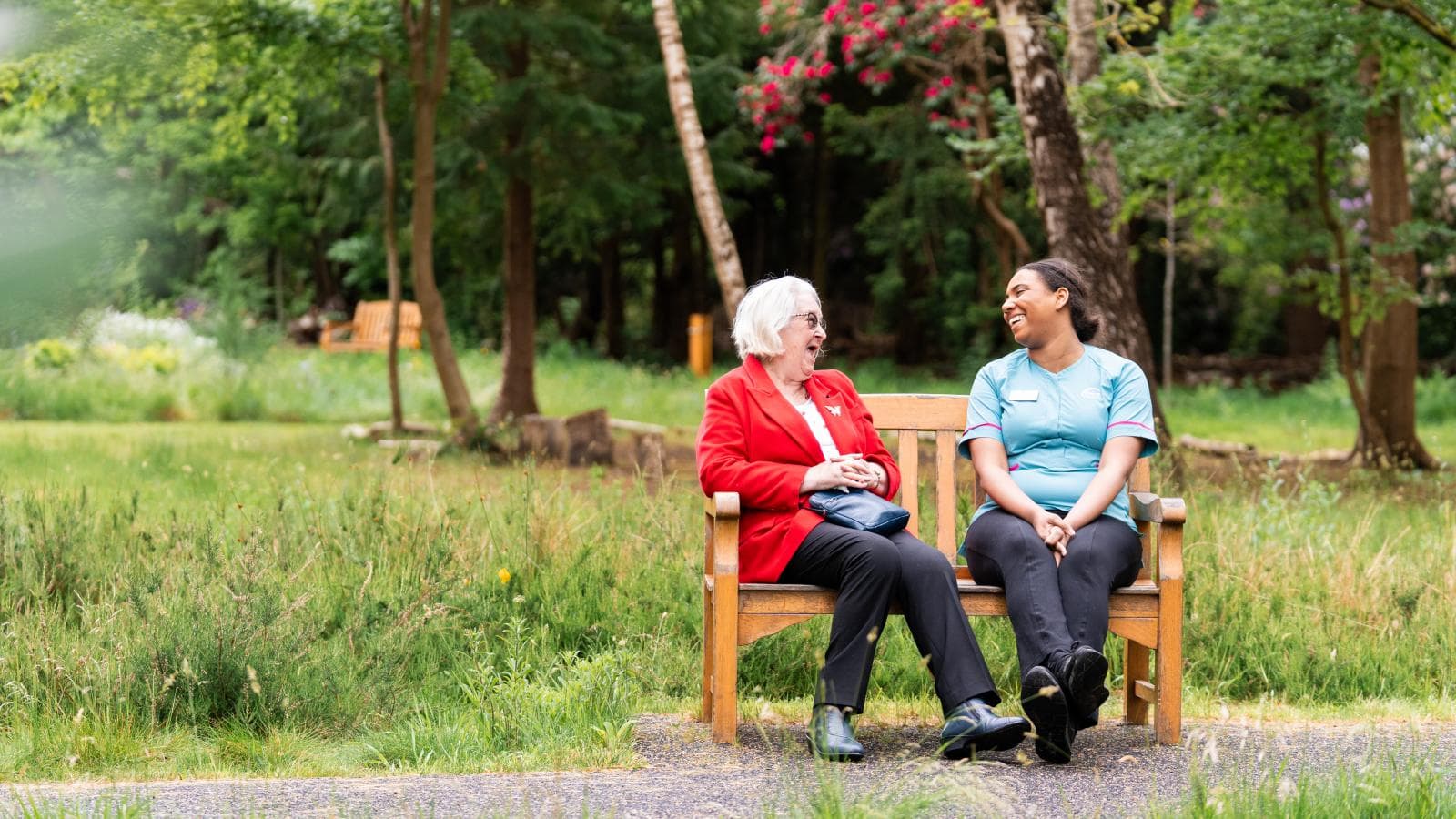 Two women sat on a garden bench, one is a female care assistant and the other is an older lady wearing a red jacket
