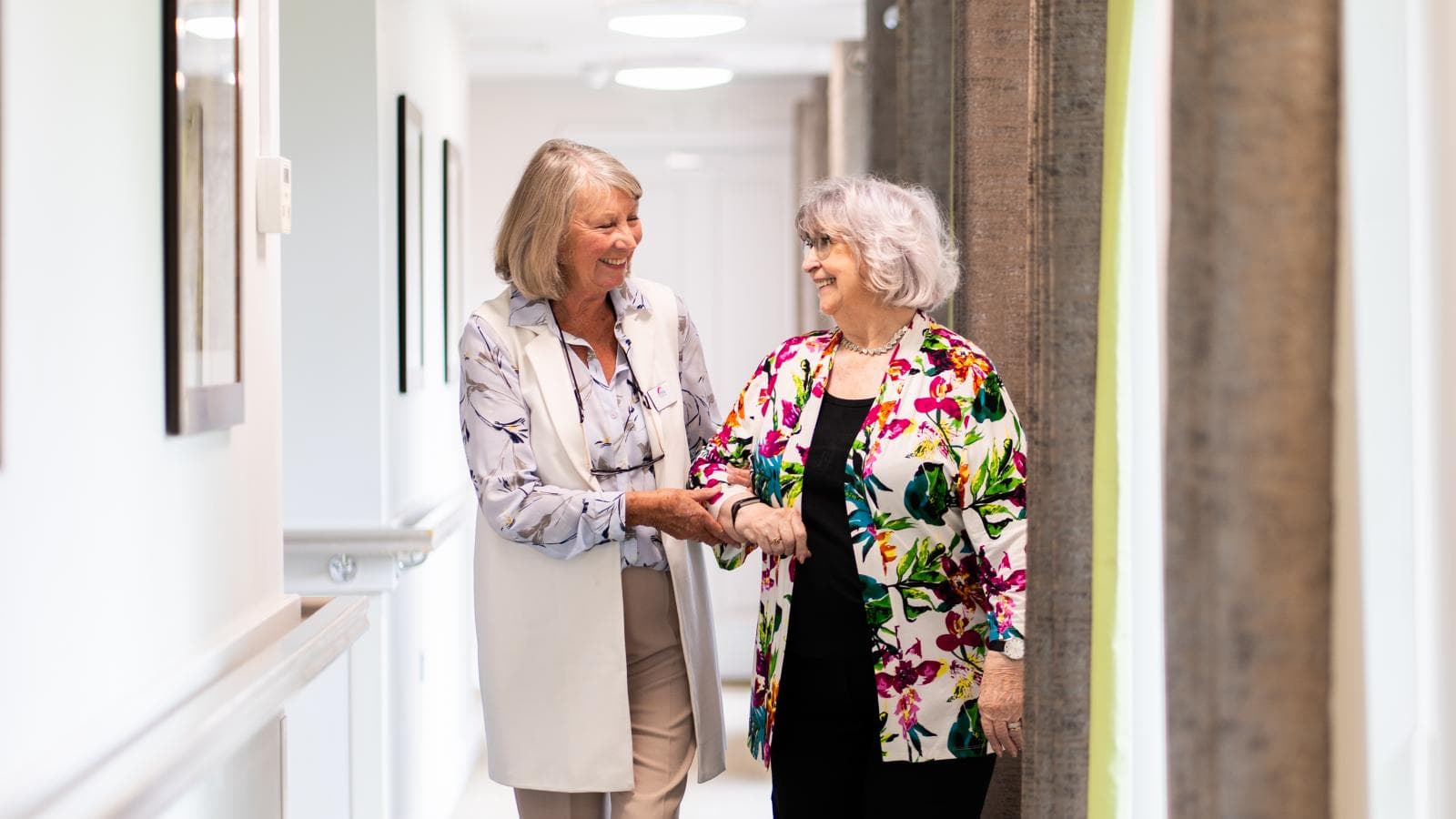 Two older ladies linking arms walking down a corridor