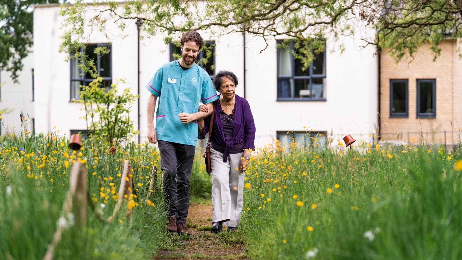 Male care assistant linking arms with elderly female care home resident walking down a garden path