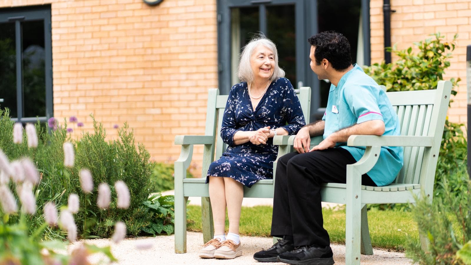 Female care home resident sat on a garden bench speaking to a male care assistant