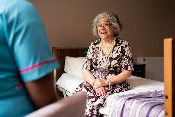 Older female care home resident sat on bed speaking to a care assistant