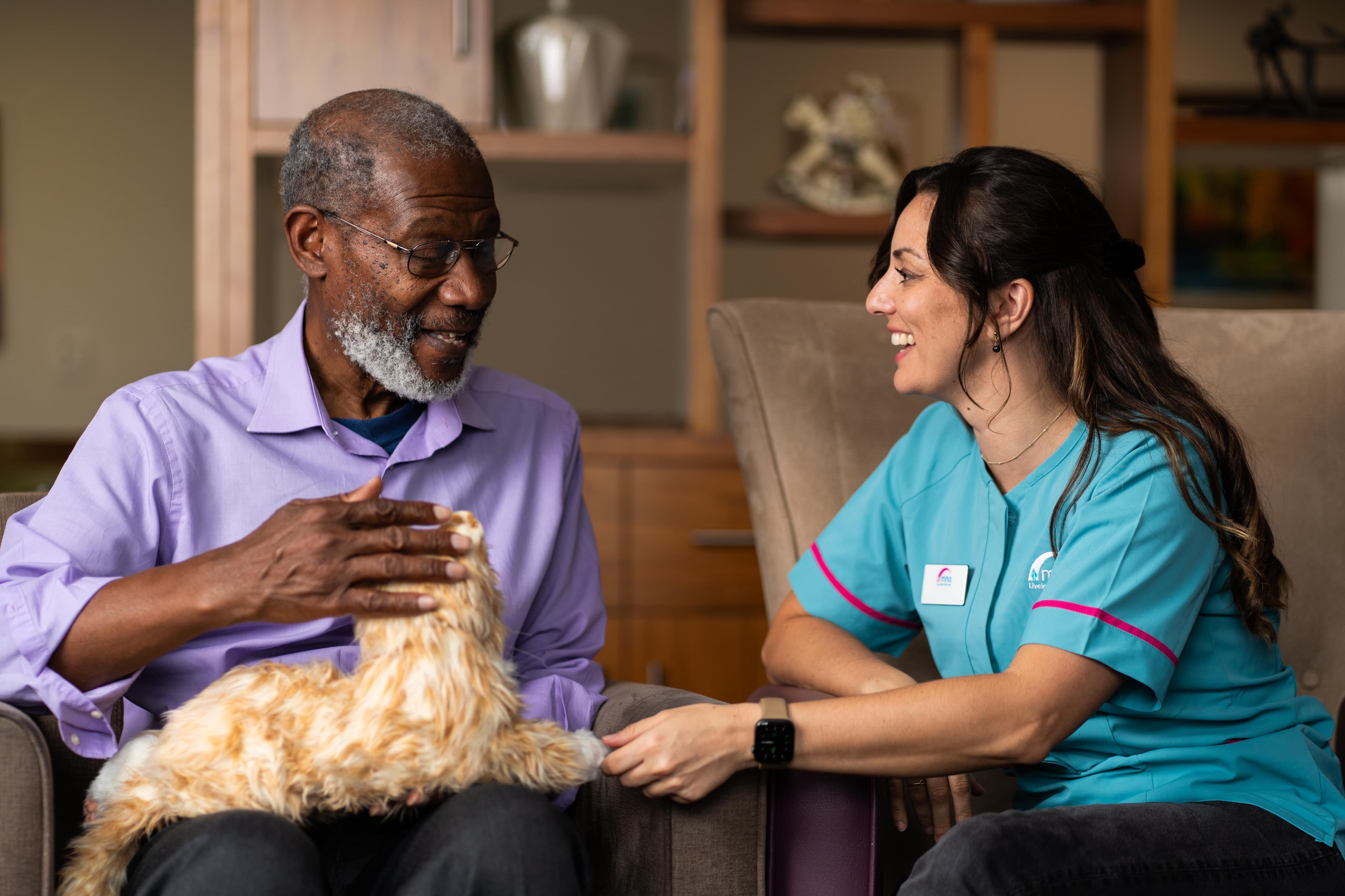 Older male with grey hair stroking robotic cat whilst sat with a female care assistant