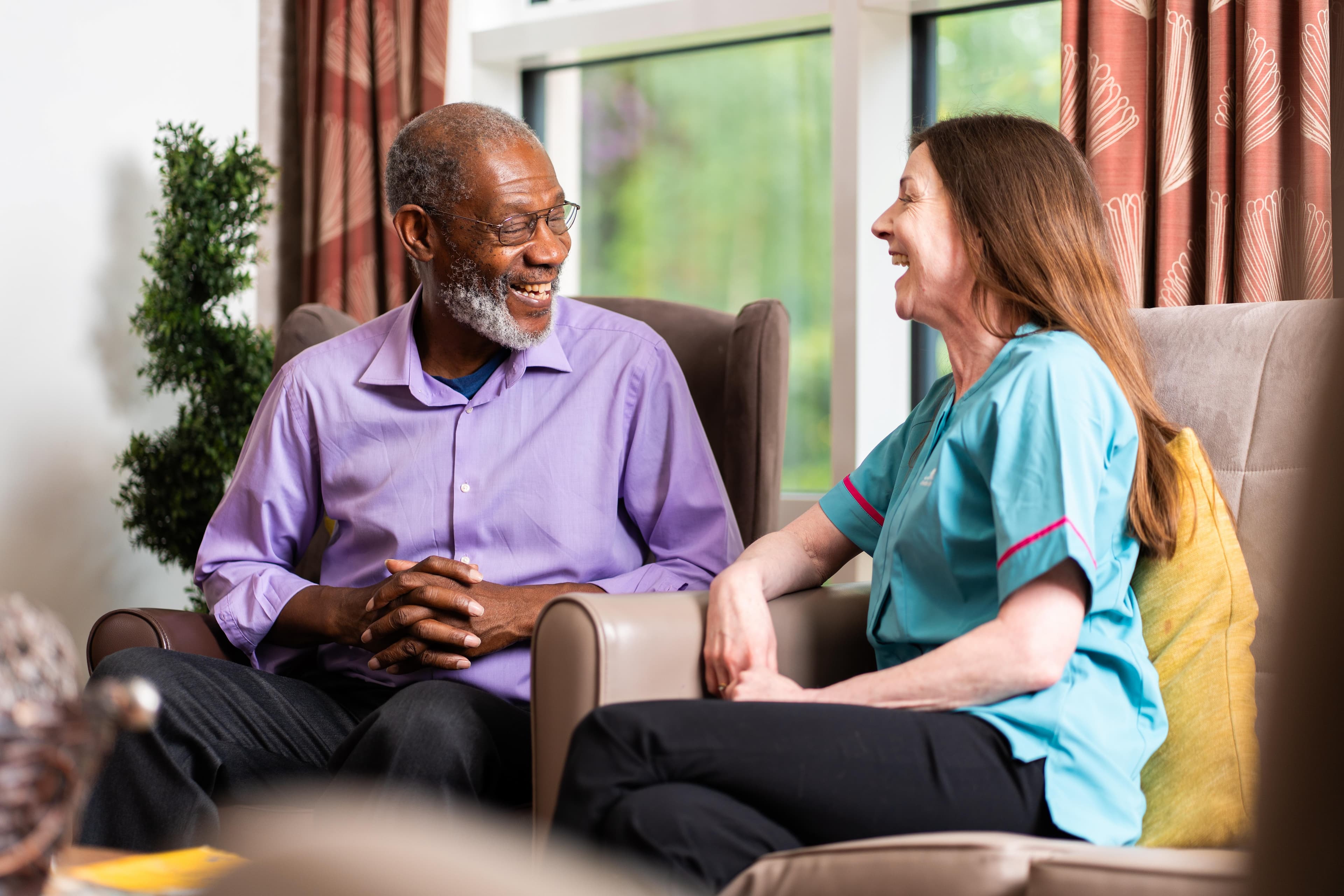 Older man with grey hair wearing a purple shirt and sat in an armchair laughing with female care assistant